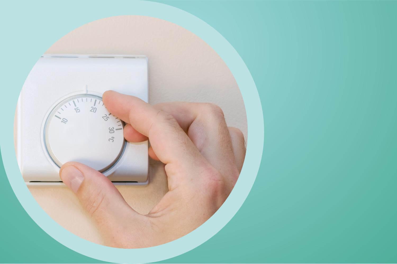 A hand turns the dial on a thermostat, mounted to a beige wall.