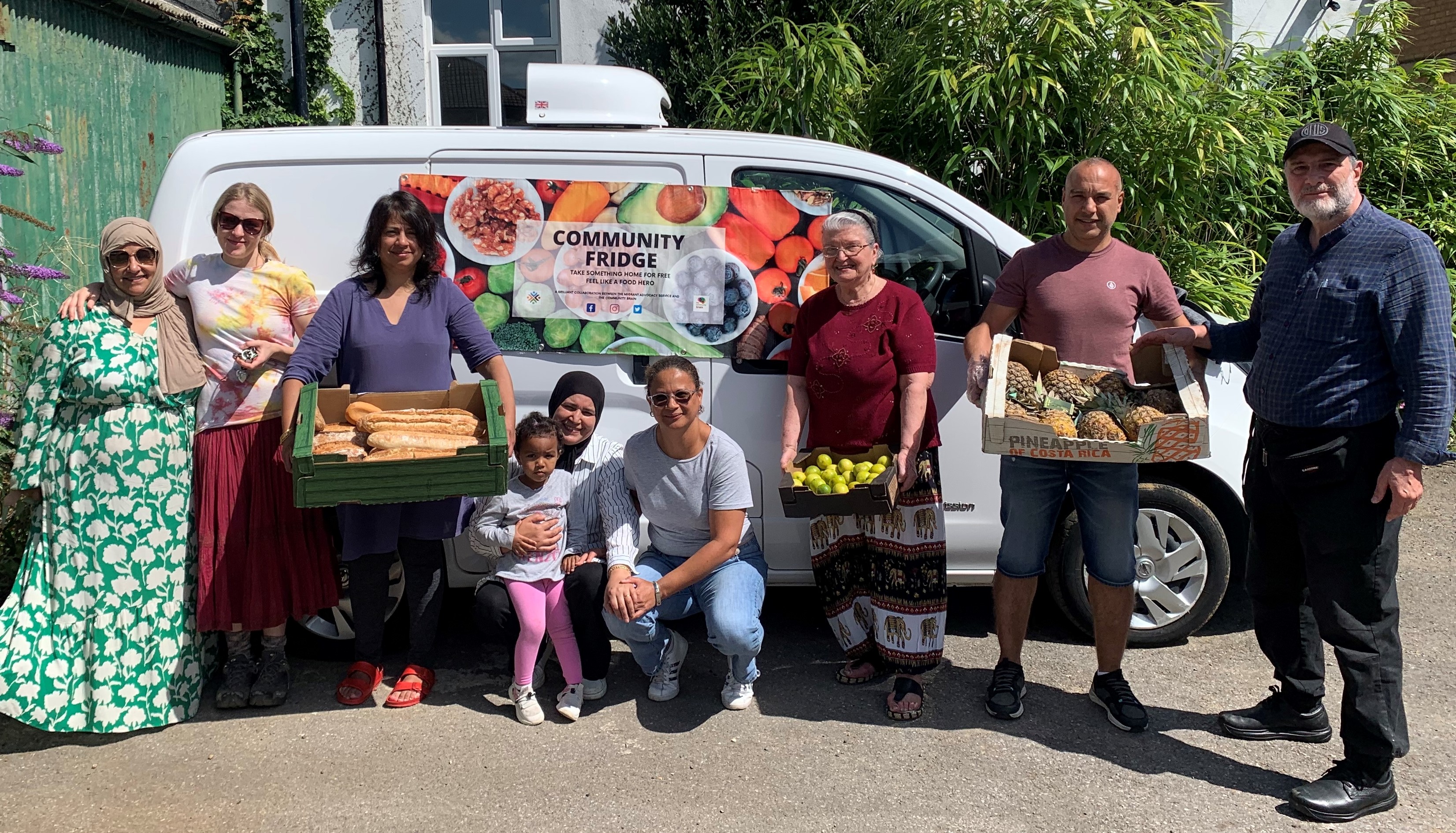 Photo of 8 voluteers from the Migrant Advisory Service holding boxes of french bread, lemons and pineapples next to their new refrigerated van and the Community Fridge banner.