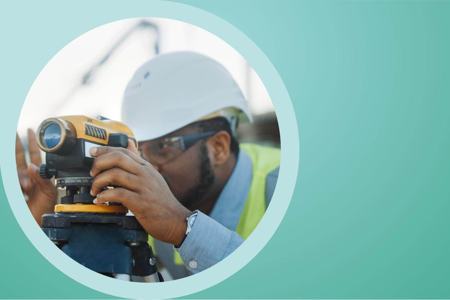 A man wearing a white hard hat and a luminous green jacket looks through a surveyors tool.