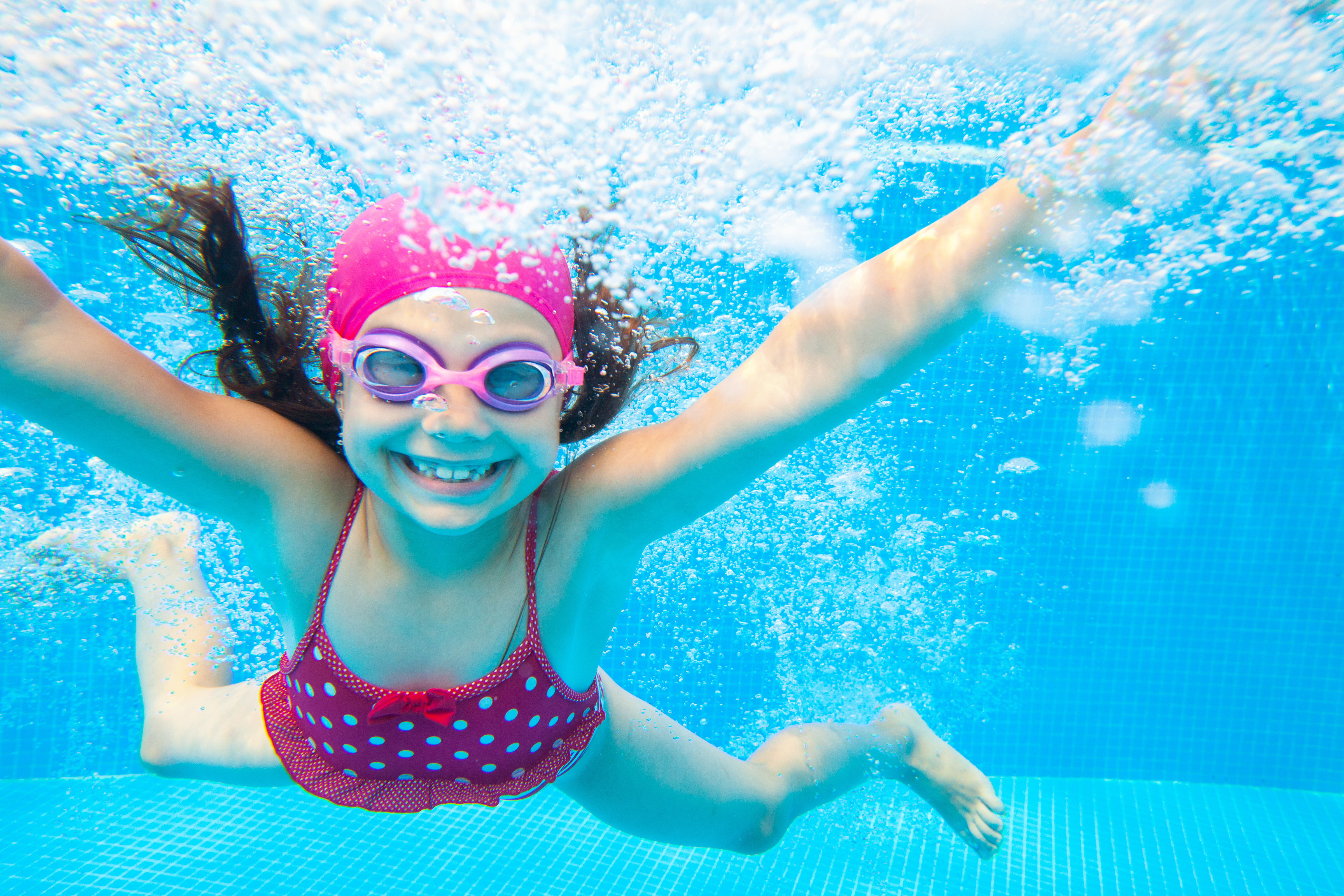 Image showing a girl underwater, smiling at the camera, wearing a pink swimming costume and swimming cap.