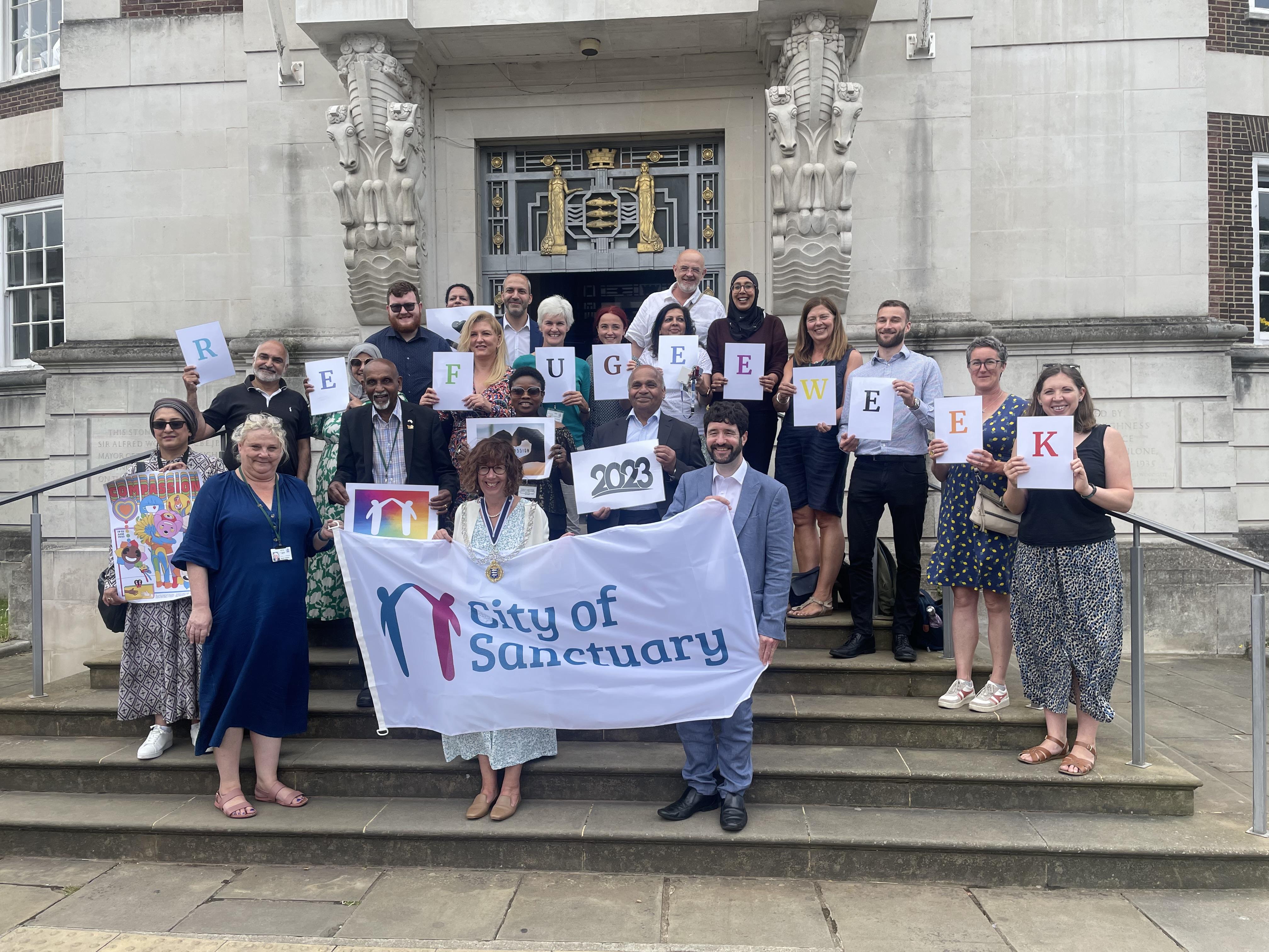 Kingston council staff standing outside guildhall holding City of sanctuary logo and letters spelling out refugee week