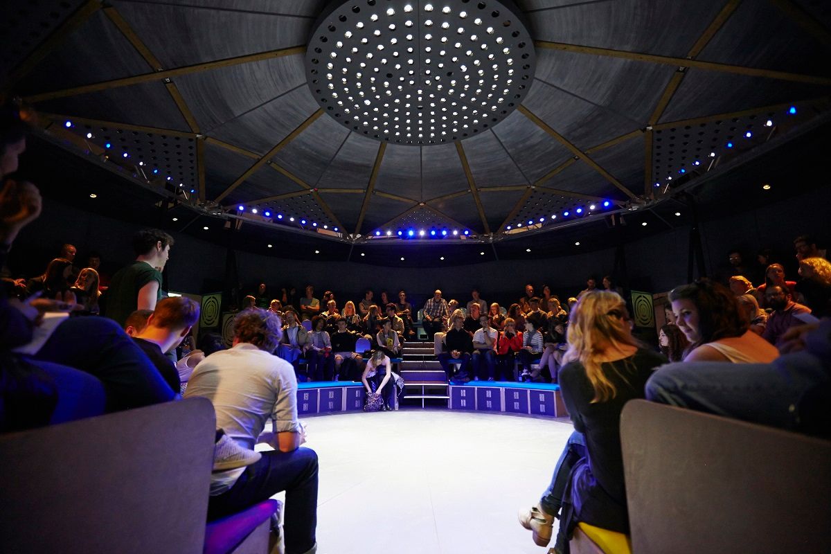 A picture of the interior of the Roundabout pop-up theatre with an audience sitting around an empty stage
