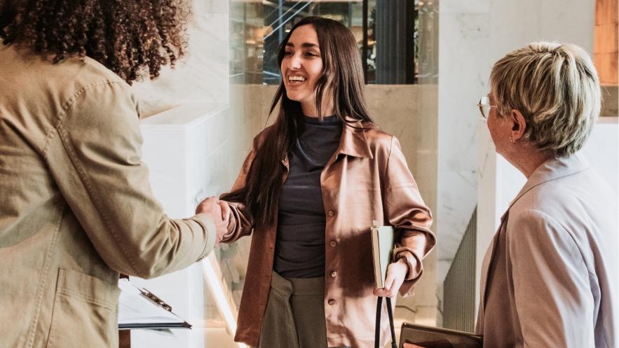 A smiling woman with long hair shakes hands with someone whose face we can't see. Another woman looks on. They are in an office setting.