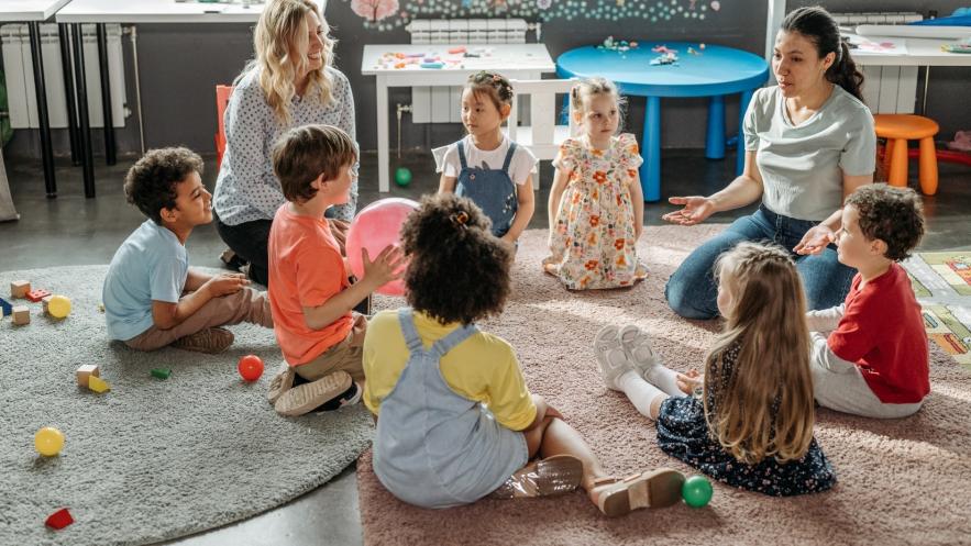 A group of pre-school children sit on the floor with two female teachers in a classroom setting, toys scattered around them