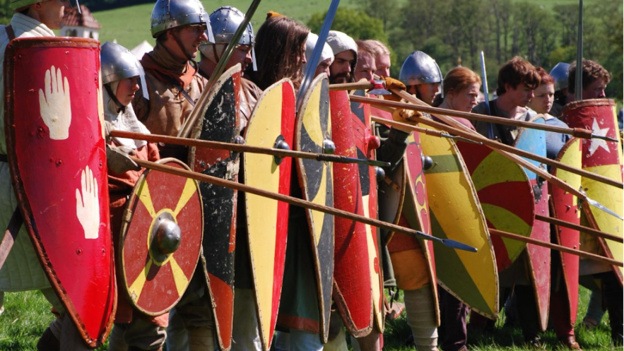 a row of warriors from the saxon era in period costume carrying large shields and spears 