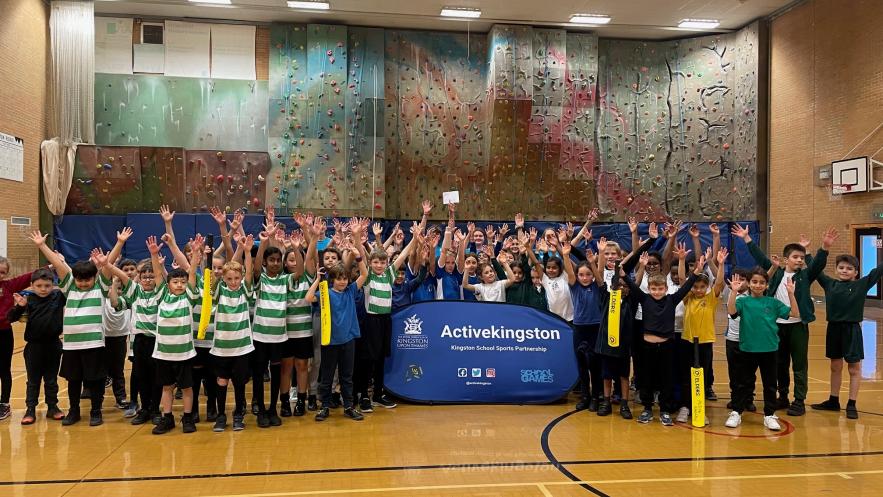 Image shows a group of children wearing sports clothes. They are standing in a sports hall, in front of a climbing wall, surrounding a blue sign that reads Active Kingston. 