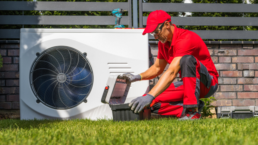 A man wearing a red hat, tshirt and trousers services a heat pump. The heat pump has a white rectangular show with a dark, greay fan to the left.